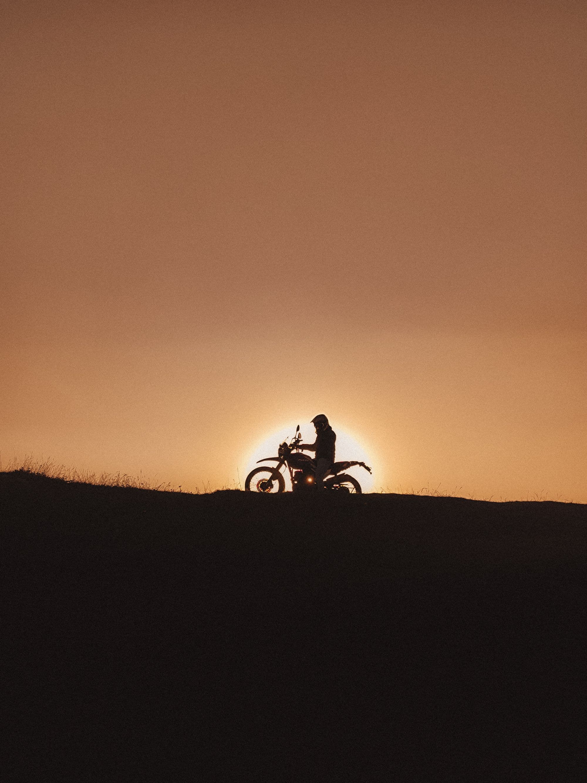 Silhouette of a rider pausing on a ridge, the setting sun glowing directly behind the motorbike